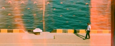 Man walks along a sunny waterfront with teal water.