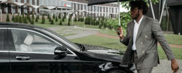 African American man in a suit walking by a car under an umbrella on a rainy day.