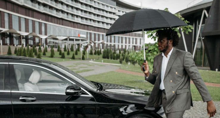 African American man in a suit walking by a car under an umbrella on a rainy day.