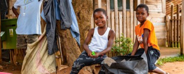 Two smiling children in front of a wooden house.
